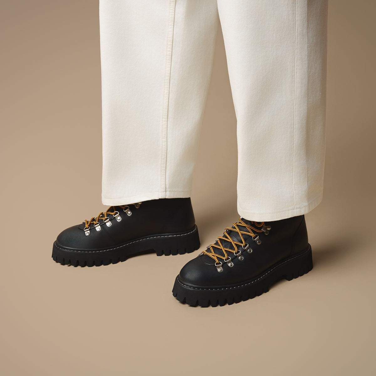 Below the knee view of man with off-white trousers wearing Blackheath black leather hiking style boots with yellow laces and d-rings on beige background