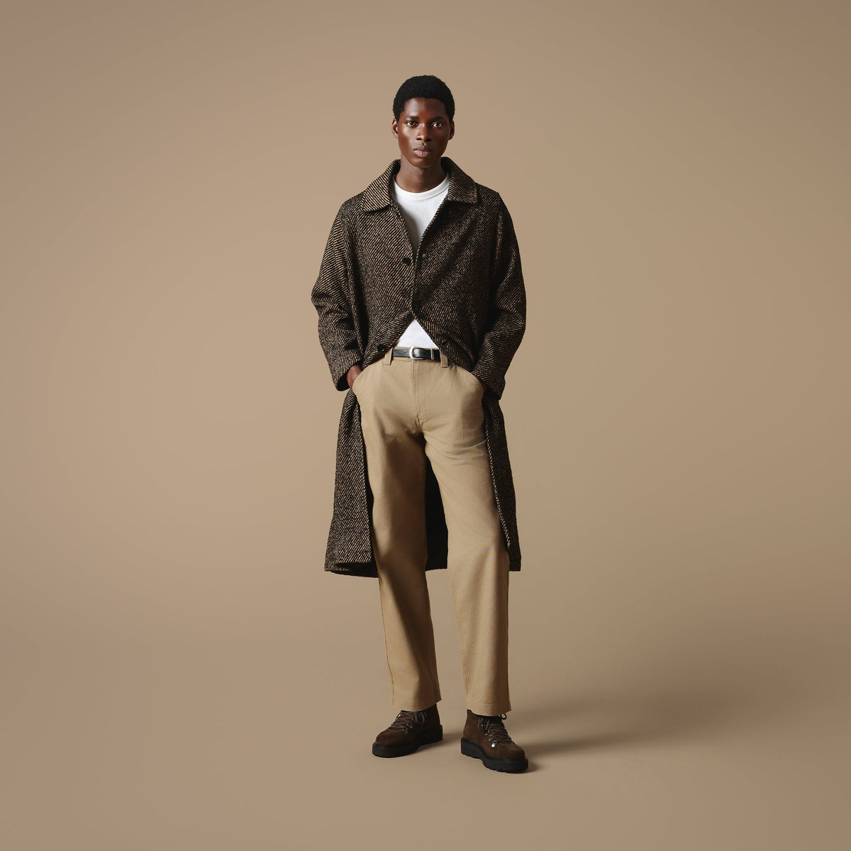 A man stood against a beige background wearing a brown overcoat, white t-shirt, paired with beige trousers and Clapham brown suede lace boots.