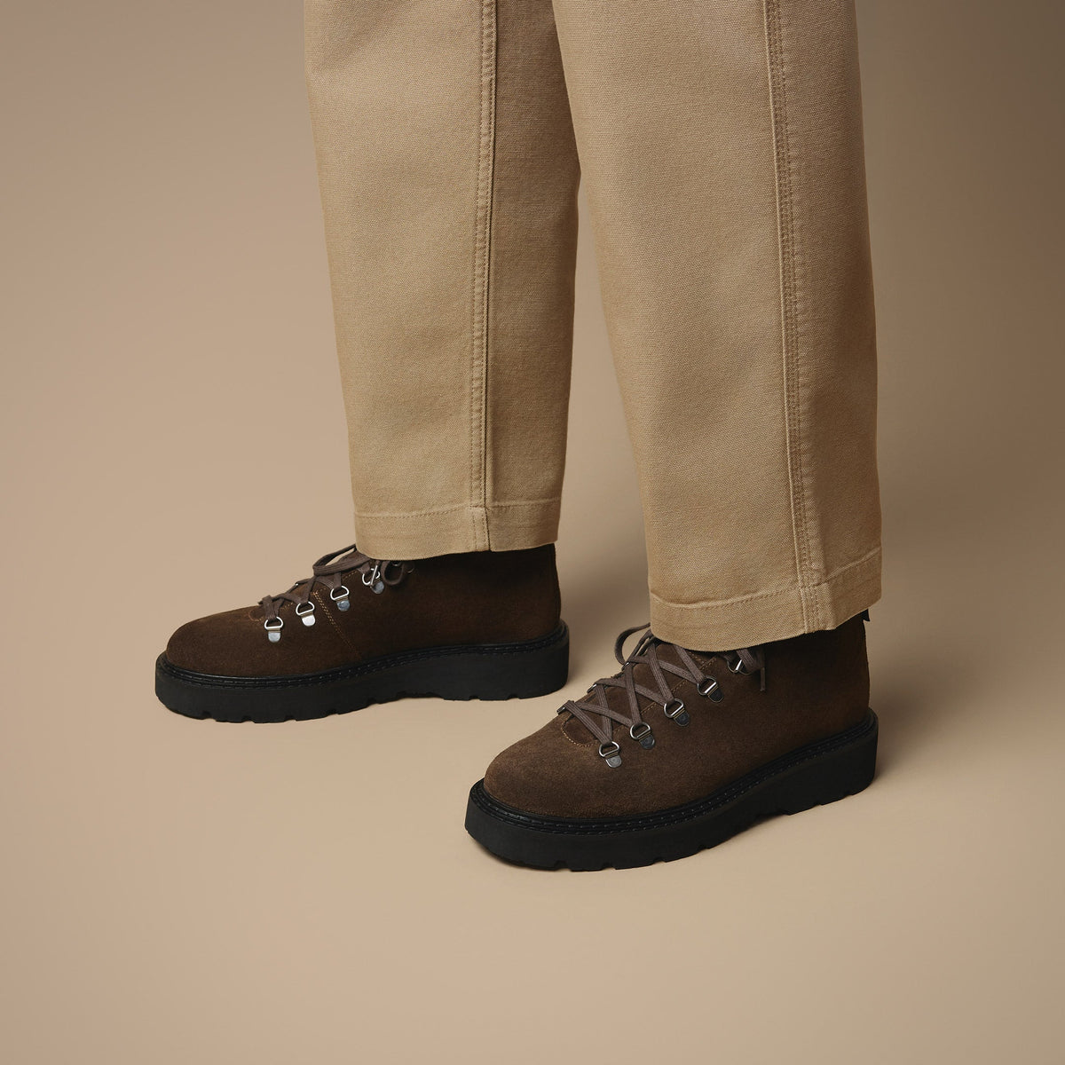 Below the knee view of man wearing beige trousers with Clapham brown suede lace boots on beige background.