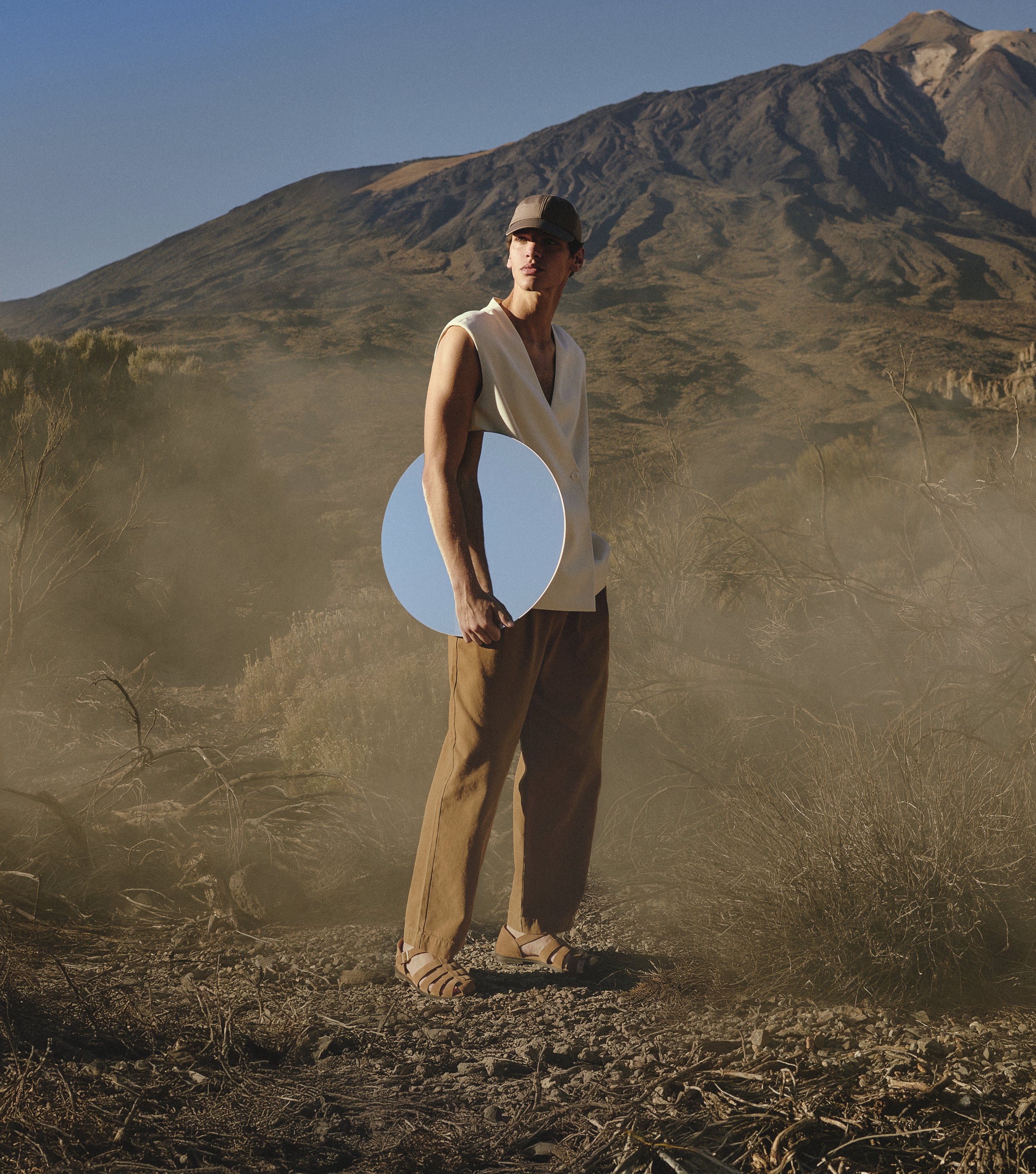 Man stood holding a circular mirror in rocky landscape wearing Promenade Sandals in Tan Suede