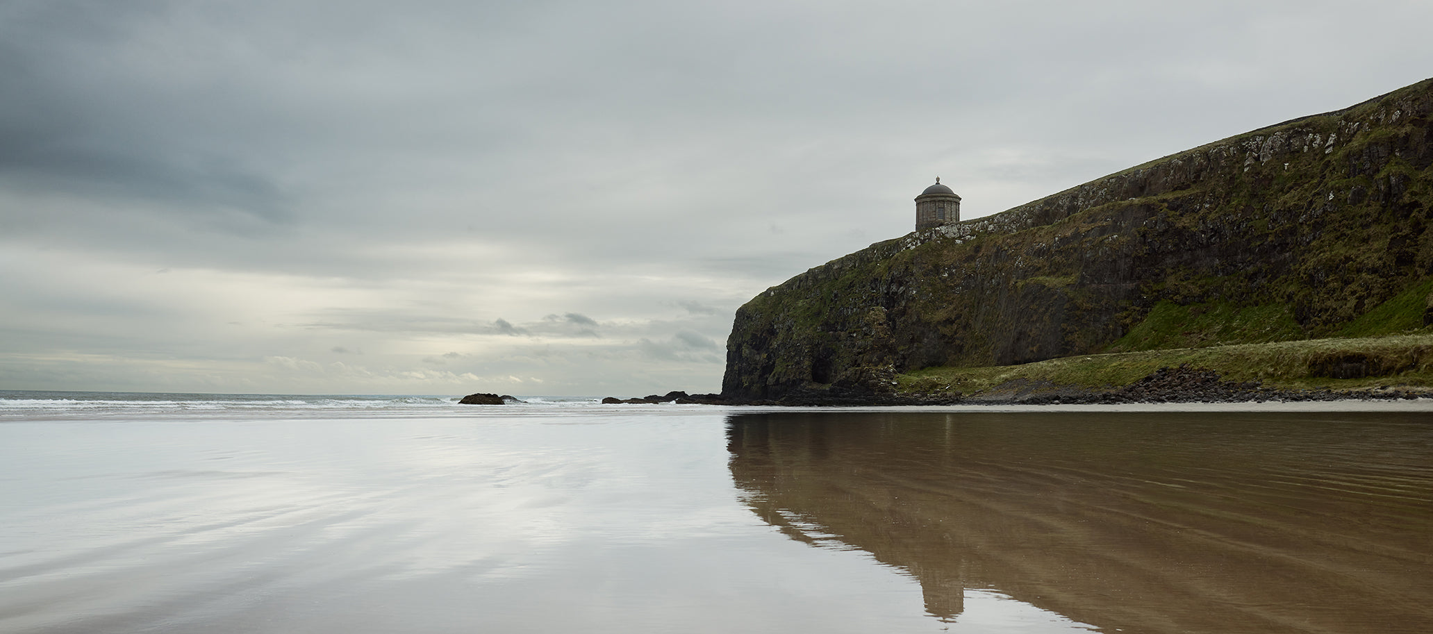 A dramatic coastal landscape with a sandy beach in the foreground reflecting the cloudy sky, a grassy cliff on the right, and a small observatory near the top of the cliff.
