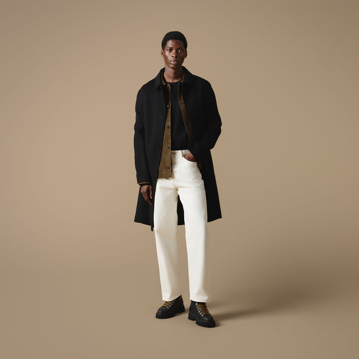 A man stood against a beige background wearing a long black coat over a brown jacket and black t-shire, paired with off-white trousers and Blackheath black leather hiking style boots.