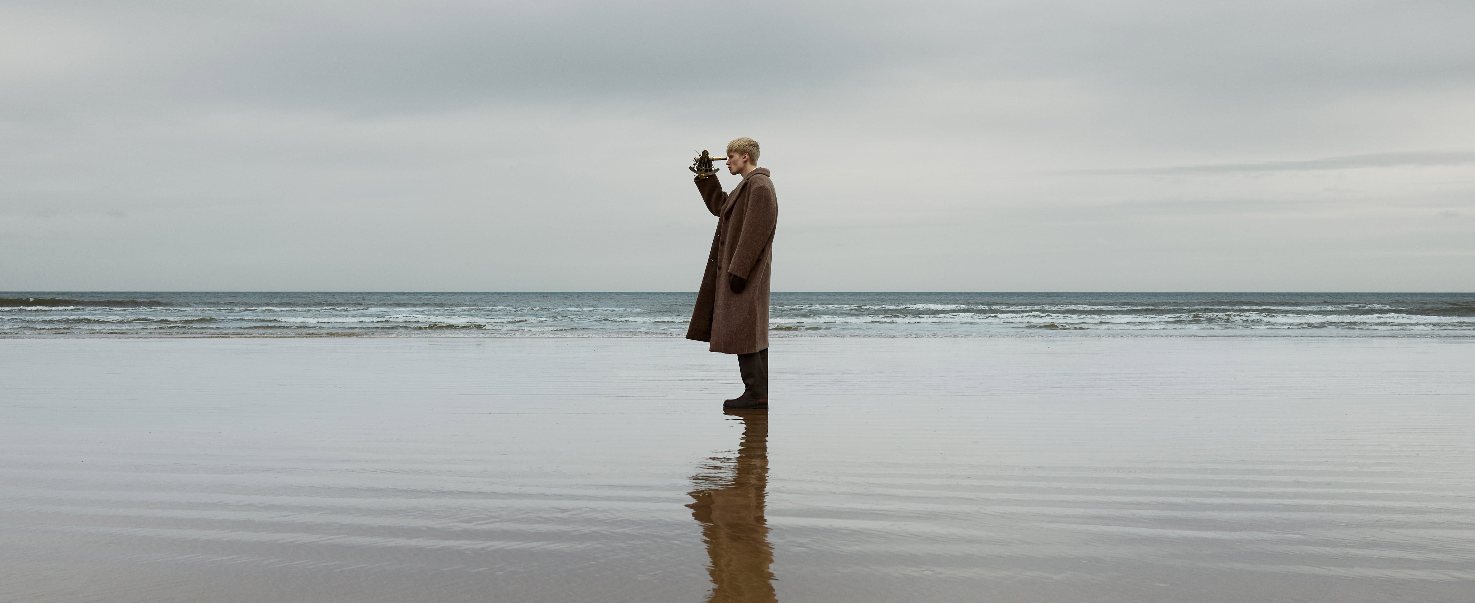 A man stood side profile on a beach on a cloudy day looking through a brass sextant and wearing long overcoat and boots