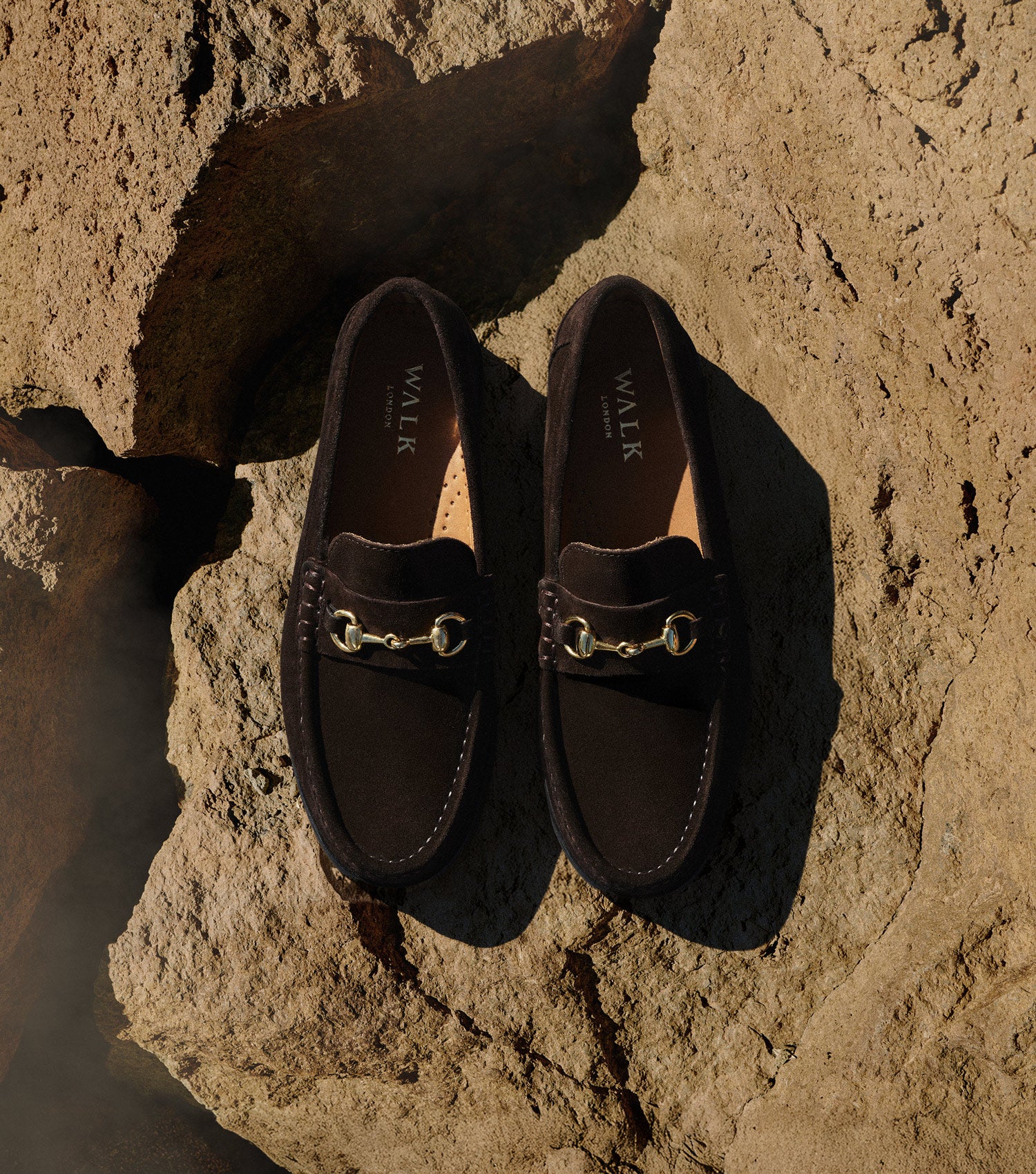 Pair of Mens Dalston Brown Suede Loafers with gold horse bit trim on rocky background