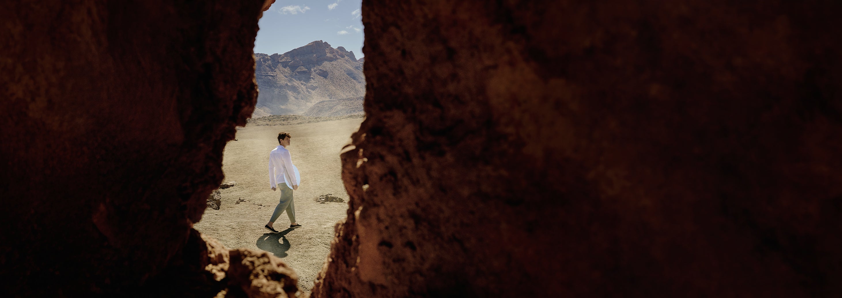 A man walking in a volcanic landscape wearing white shirt, green trousers, black slip on shoes viewed through a gap in a rock