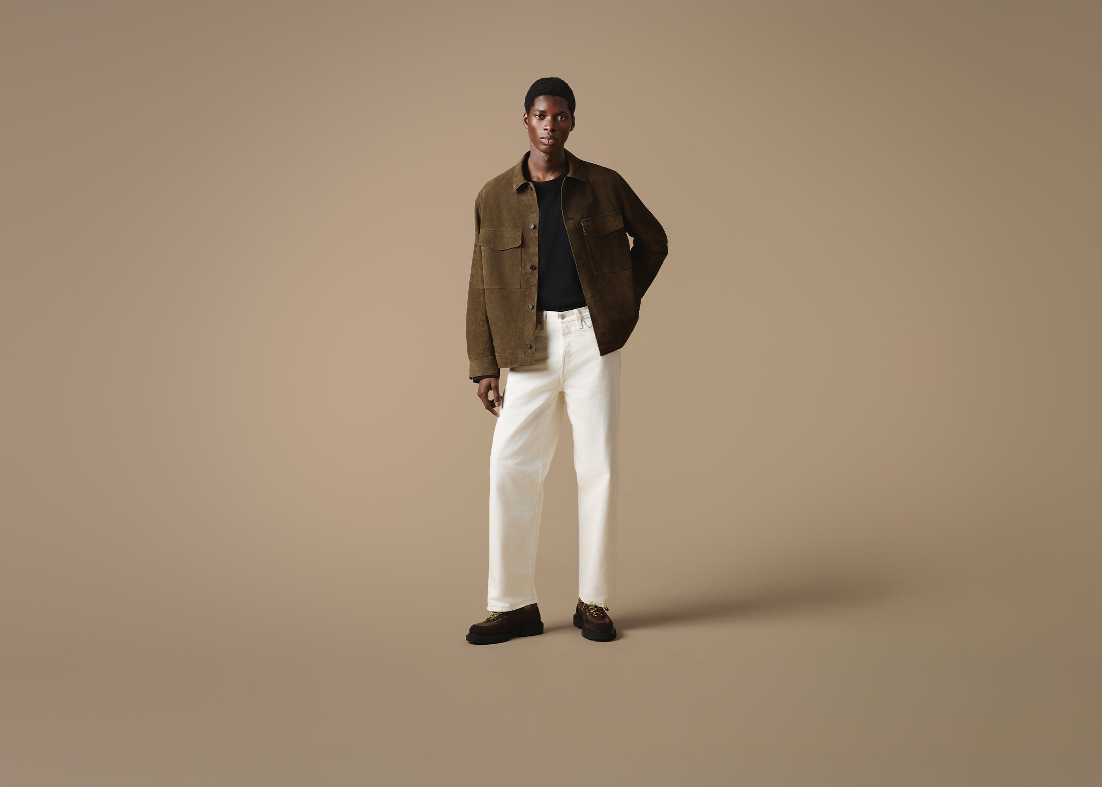 A man stood against a beige studio background wearing a brown jacket, black top, with off-white trousers and Bethnal brown leather and suede hiking style shoes