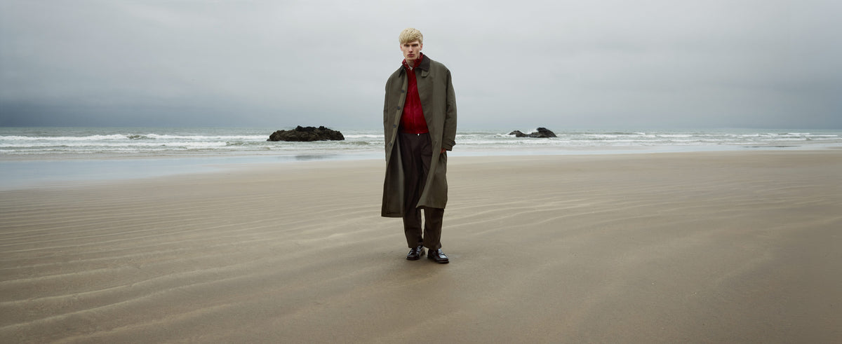 A man stood on a beach wearing long overcoat, red shirt, brown trousers and Shoreditch burgundy leather penny loafers