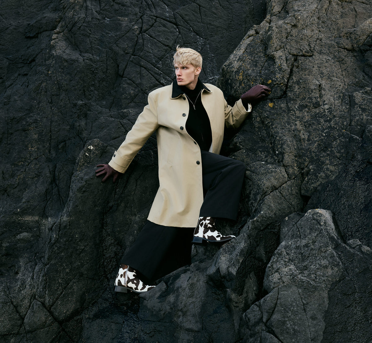 A man climbing a dark cliff wearing a long cream coat, gloves, dark trousers and Abbey cow print boots