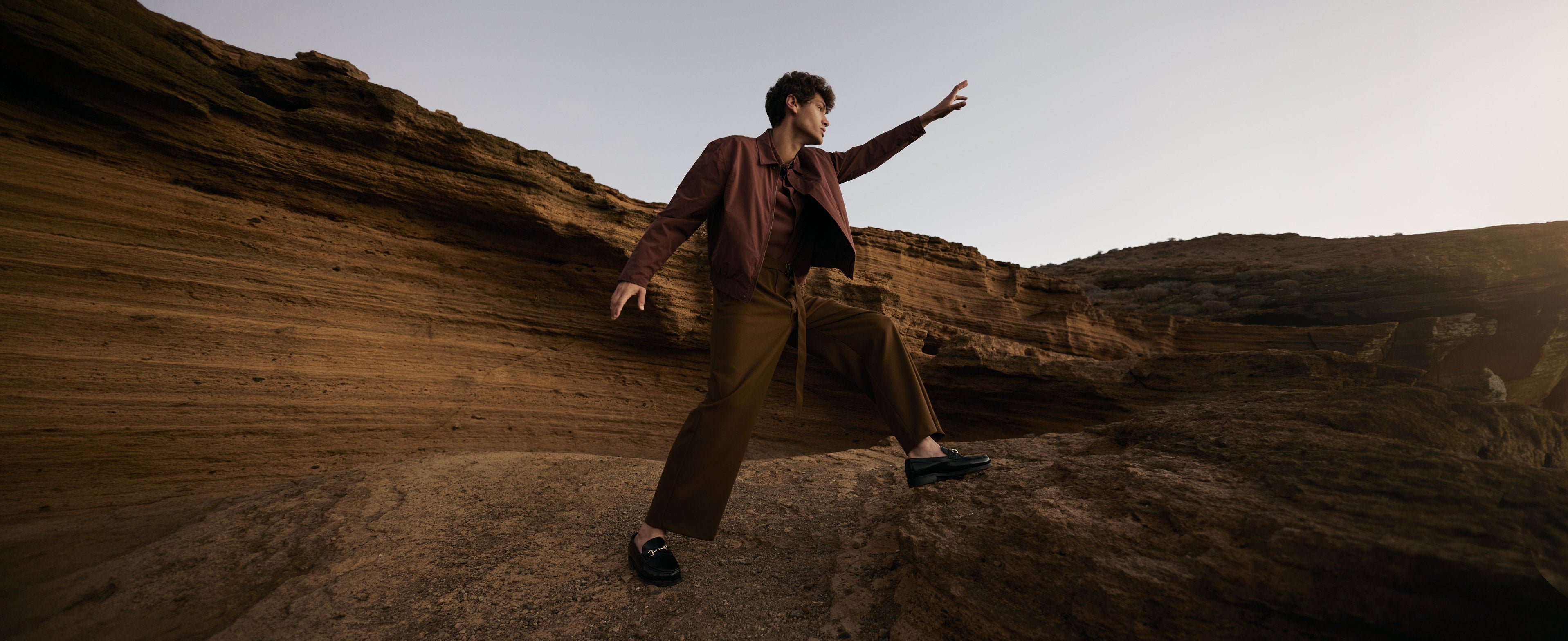 Man wearing burgundy jacket, brown trousers, brown suede trim loafers reaching for the light on rocky scene