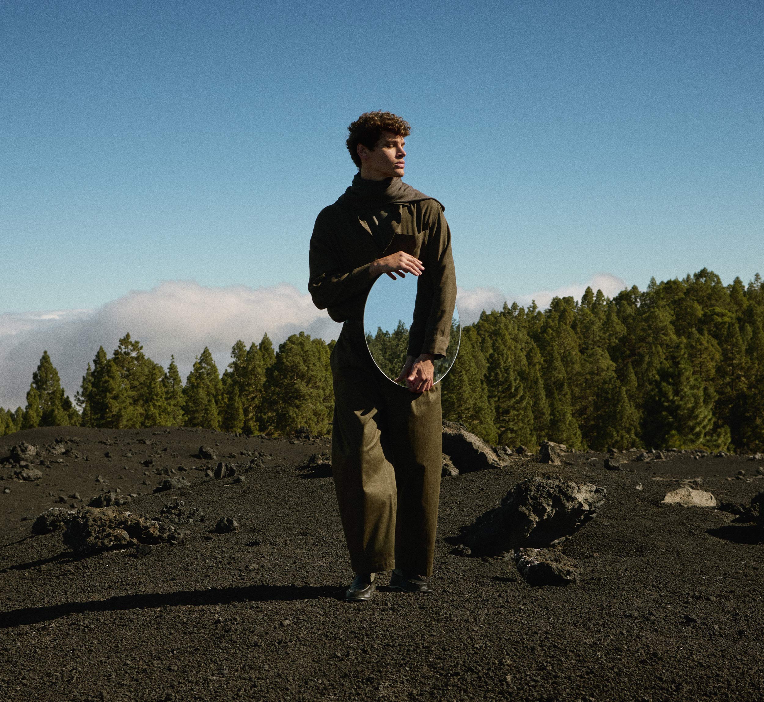 Man holding mirror walking over volcanic landscape in black loafers
