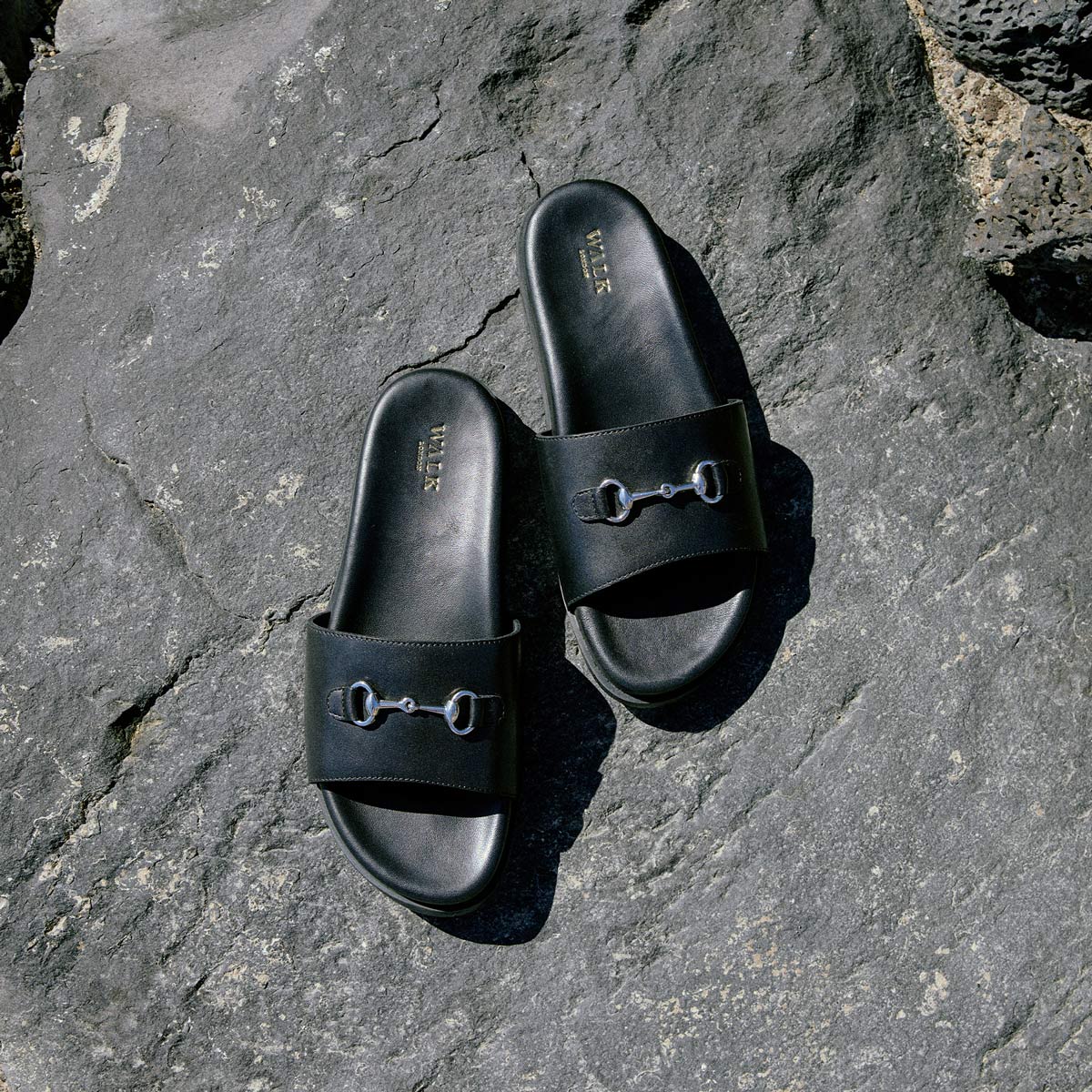 Walk London Breeze Trim Men's Black Leather Sandals with silver-toned horse bit detail on grey rock