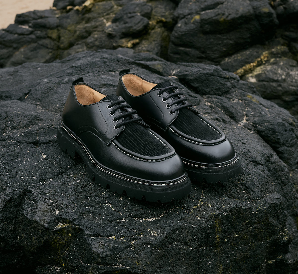 A pair of Farringdon black leather lace shoes with corded suede vamp on dark rocks on the beach.