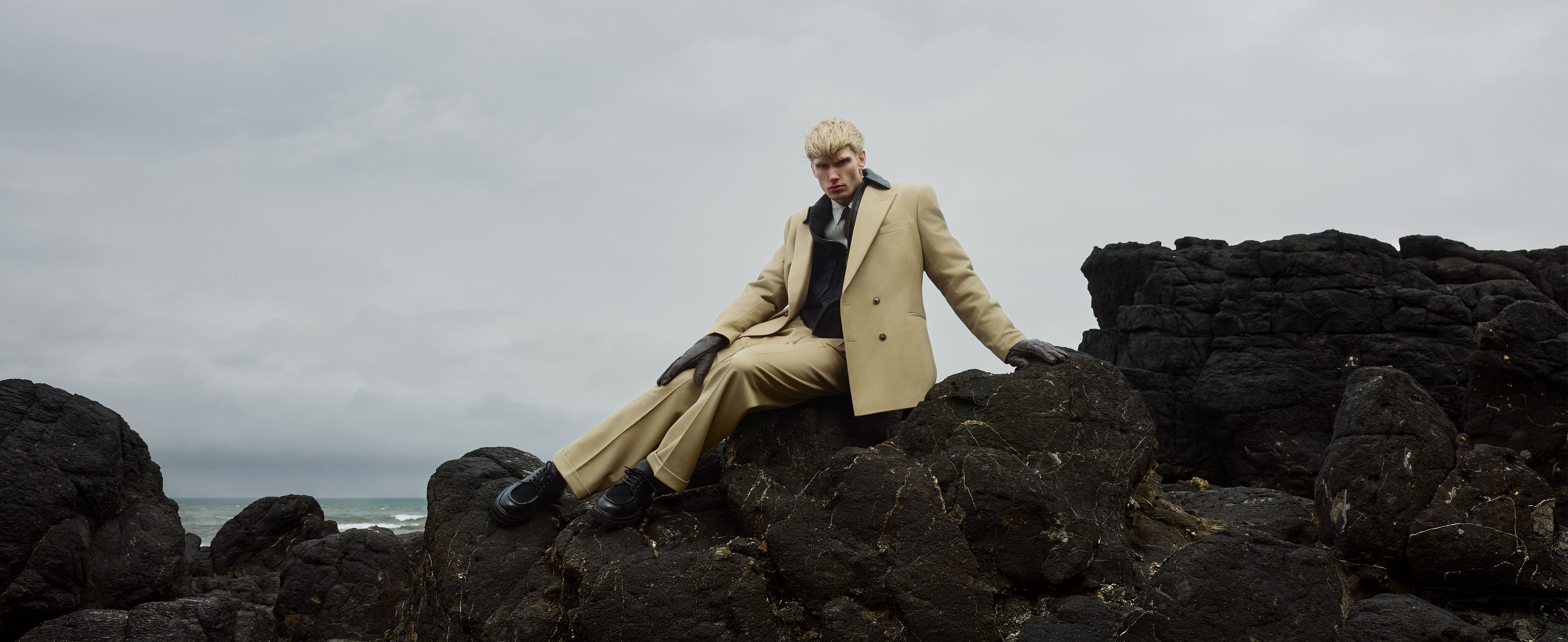 A man in a beige suit sits on black rocks by the coast wearing black leather shoes and gloves