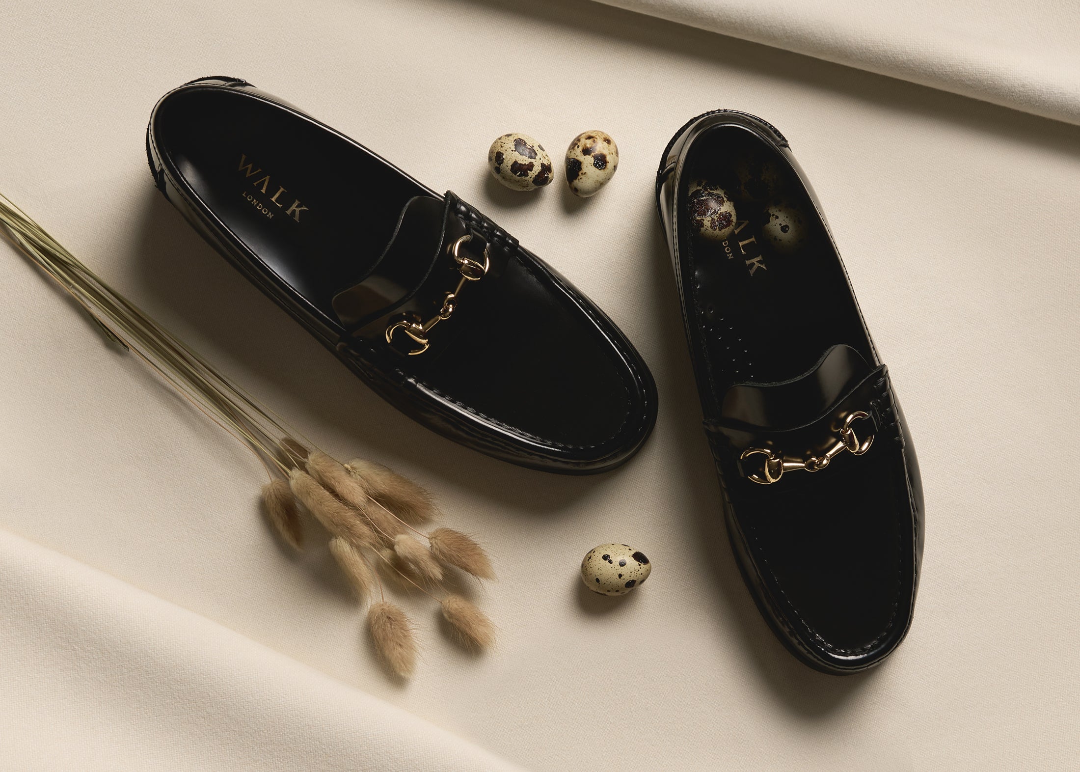A pair of Dalston black leather trim loafers on cream background with quail eggs and dried bunny tail grass