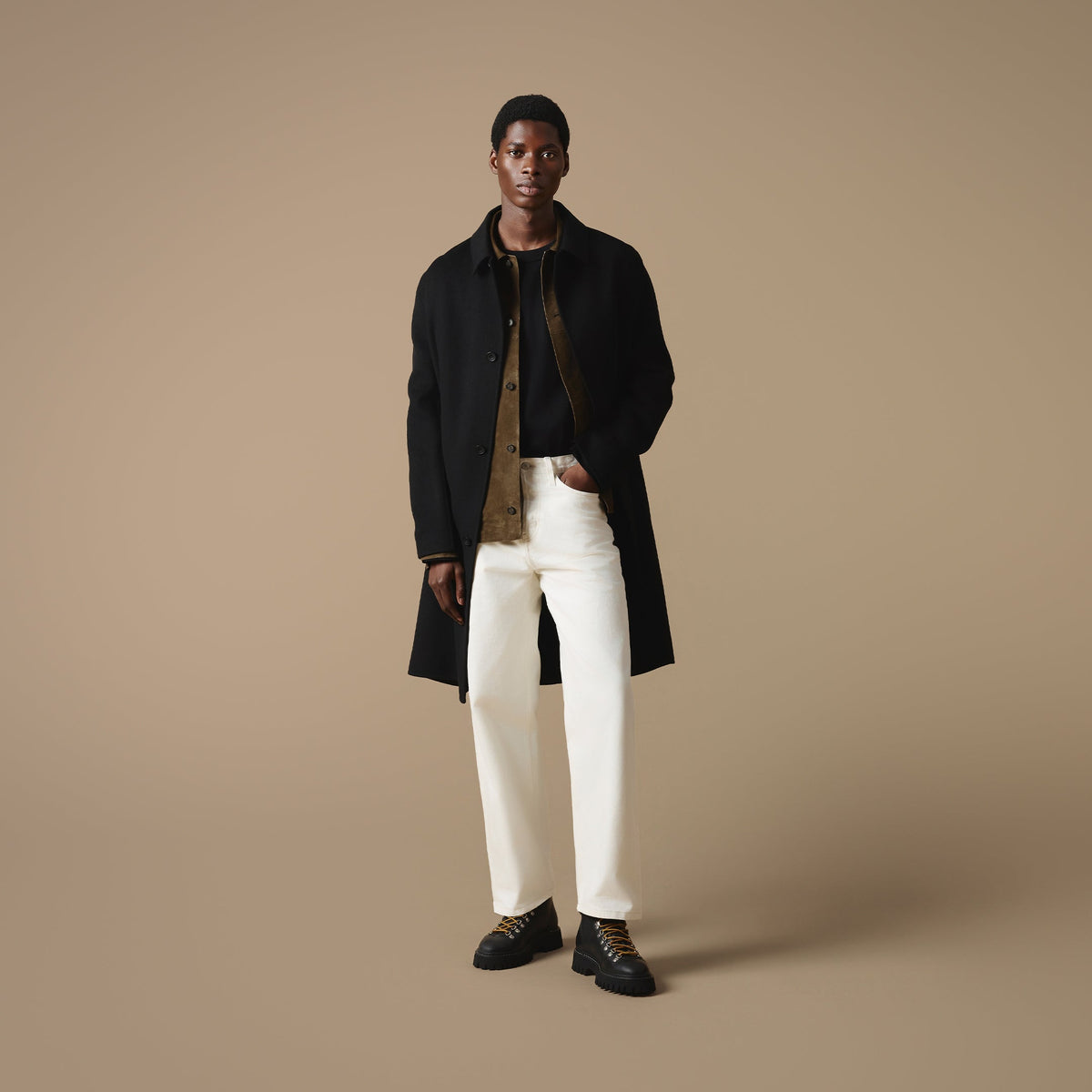 A man stood against a beige background wearing a long black coat over a brown jacket and black t-shire, paired with off-white trousers and Blackheath black leather hiking style boots.
