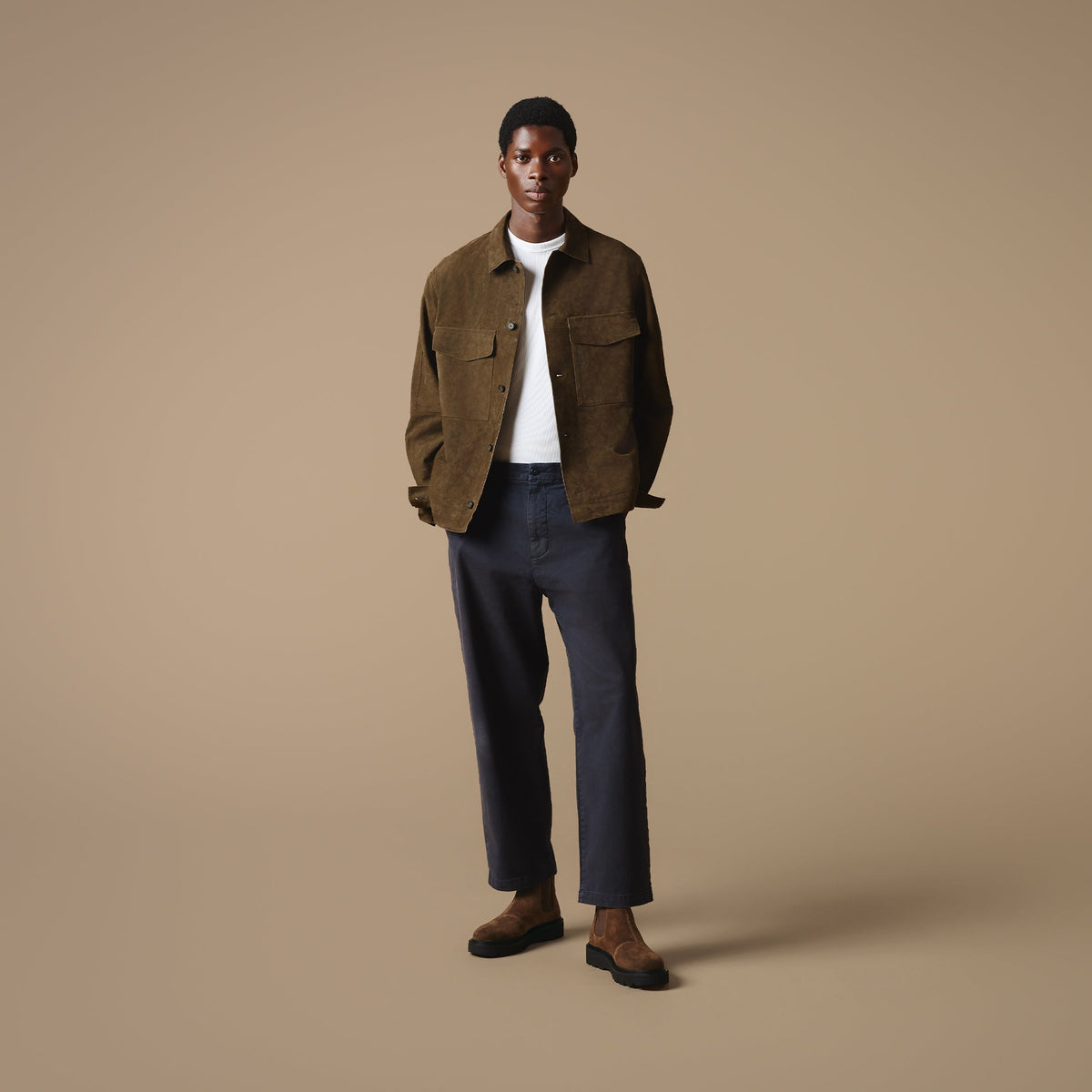 A man stood against a beige background wearing a brown jacket over a white t-shirt, dark blue jeans, and Clapham tan suede Chelsea boots.