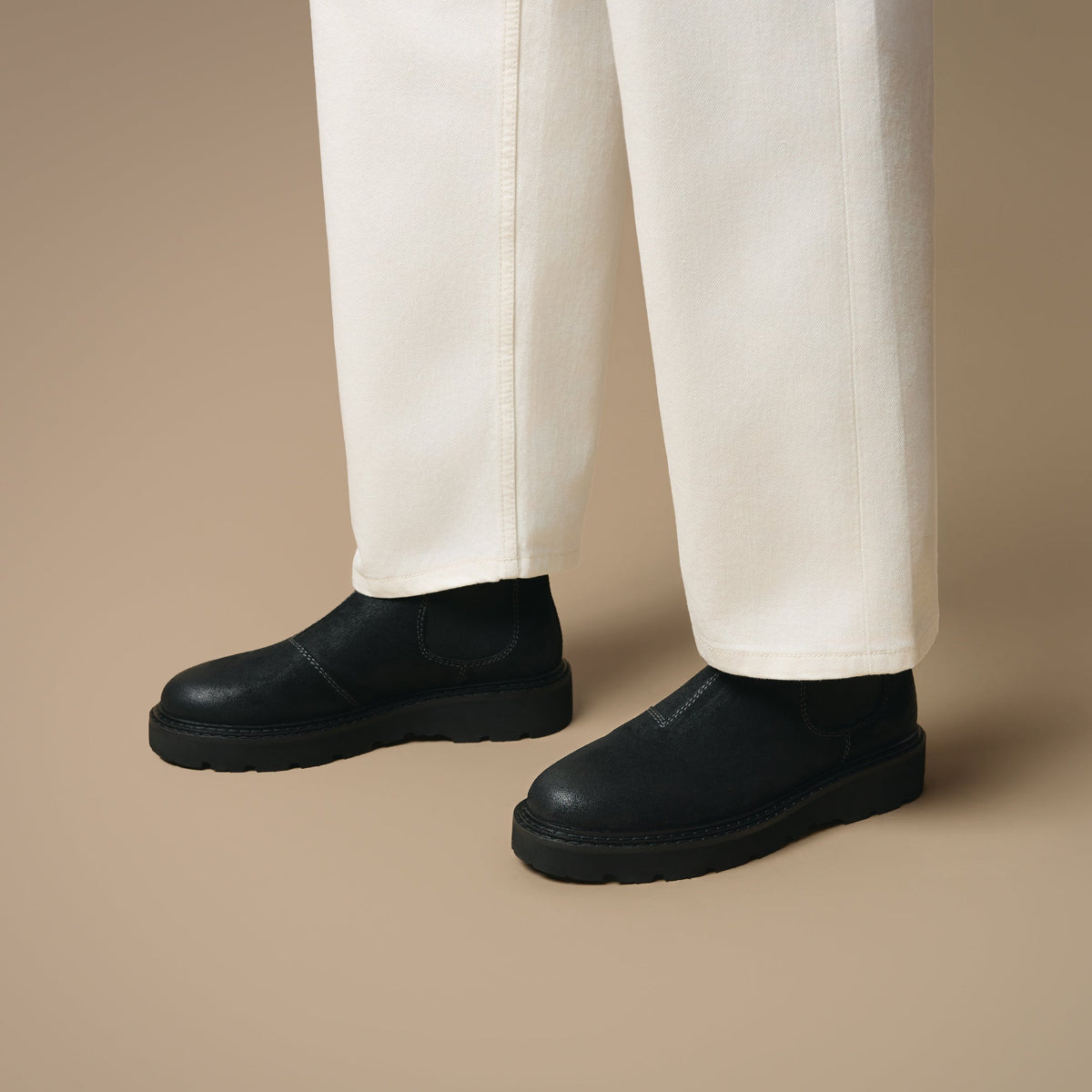 Below the knee view of man wearing off-white trousers and Clapham black suede Chelsea boots on beige background.