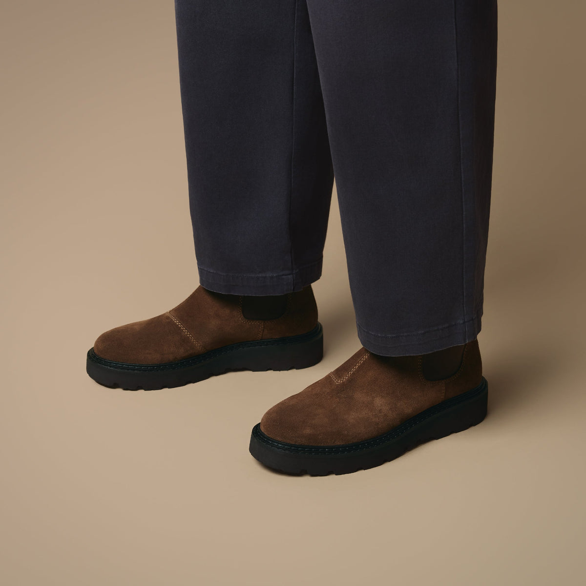 Below the knee view of man wearing dark blue jeans and Clapham tan suede Chelsea boots on beige background