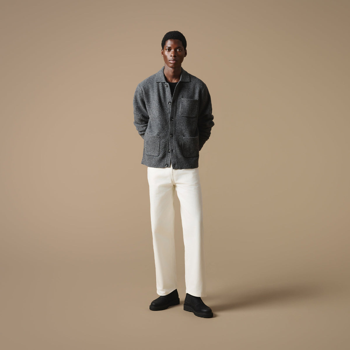 A man stood against a beige background wearing a grey textured button-up top, off-white trousers, and Clapham black suede Chelsea boots.