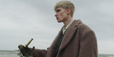 Video of blond man holding a brass sextant, walking on a beach on a cloudy day wearing hair-on leather boots