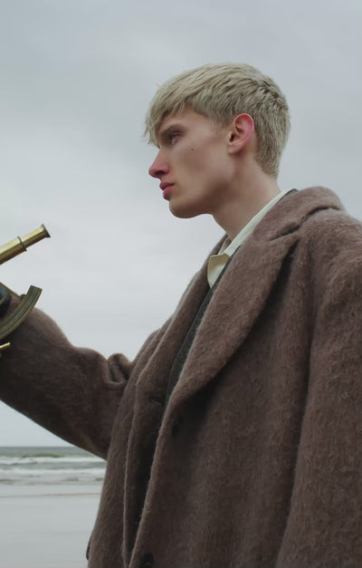 Video of blond man holding a brass sextant, walking on a beach on a cloudy day wearing hair-on leather boots